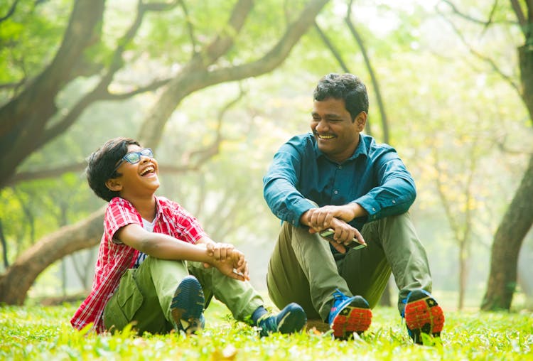 A Man And A Boy Laughing While Sitting On The Gree Grass