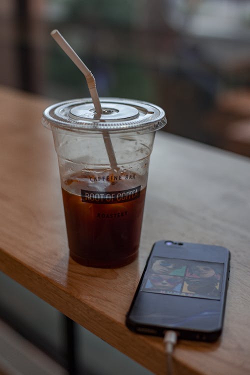 From above of cup of cold coffee with ice and straw placed on table in cafe with cellphone   on charge