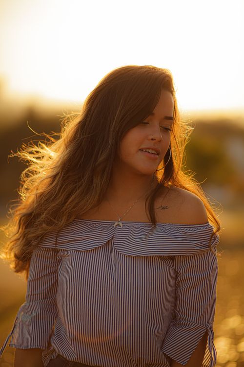 Free Pretty young female wearing blouse with bare shoulders looking away while standing in sunlight in meadow in summer evening with blurred background Stock Photo