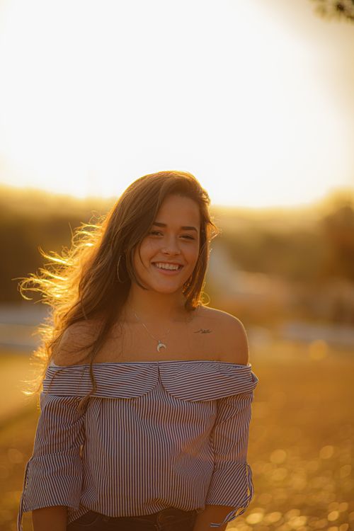 Free Happy female with long hair smiling and looking at camera while standing under sunbeams on field at evening time with blurred background Stock Photo