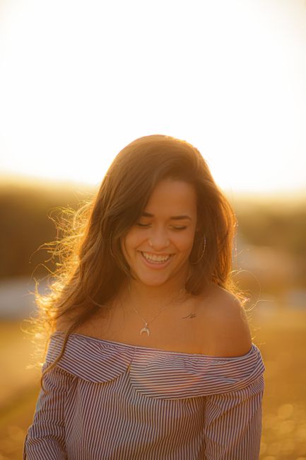 Delighted lady with eyes closed and long hair smiling while standing in sunlight in blurred rural field at sunset time
