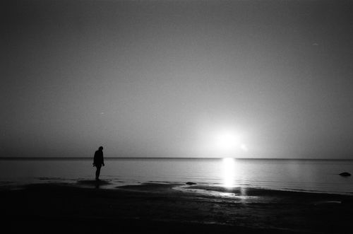 Free Silhouette of a person standing by the tranquil sea during a sunrise, captured in black and white. Stock Photo