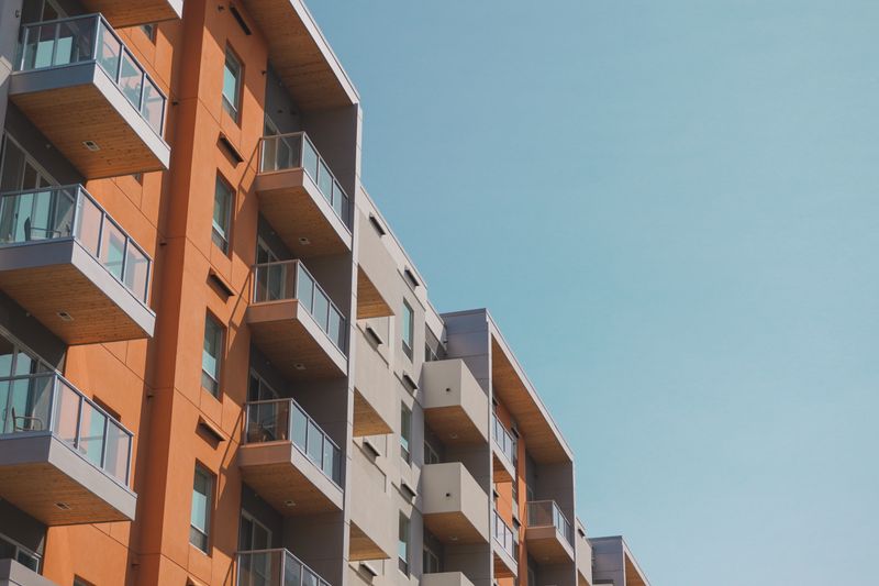 A mid-rise residential building with stacked glass-railed balconies and orange and grey panelling, against a clear sky.