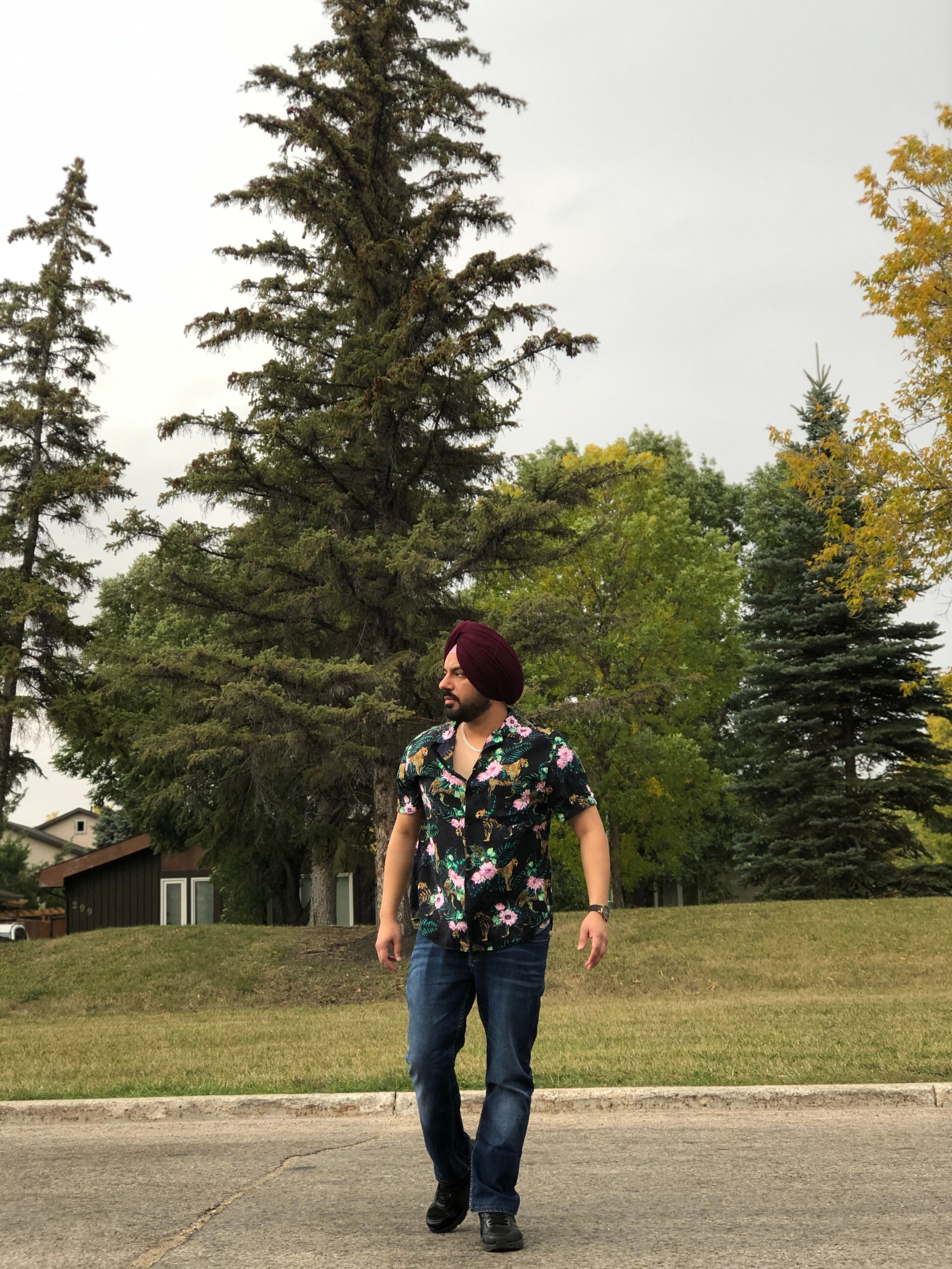Man in a floral shirt and turban walking outdoors in a scenic park with tall trees, embodying a relaxed lifestyle.
