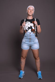 Tattooed woman in denim shorts and graphic tee against gray background, showcasing modern style.