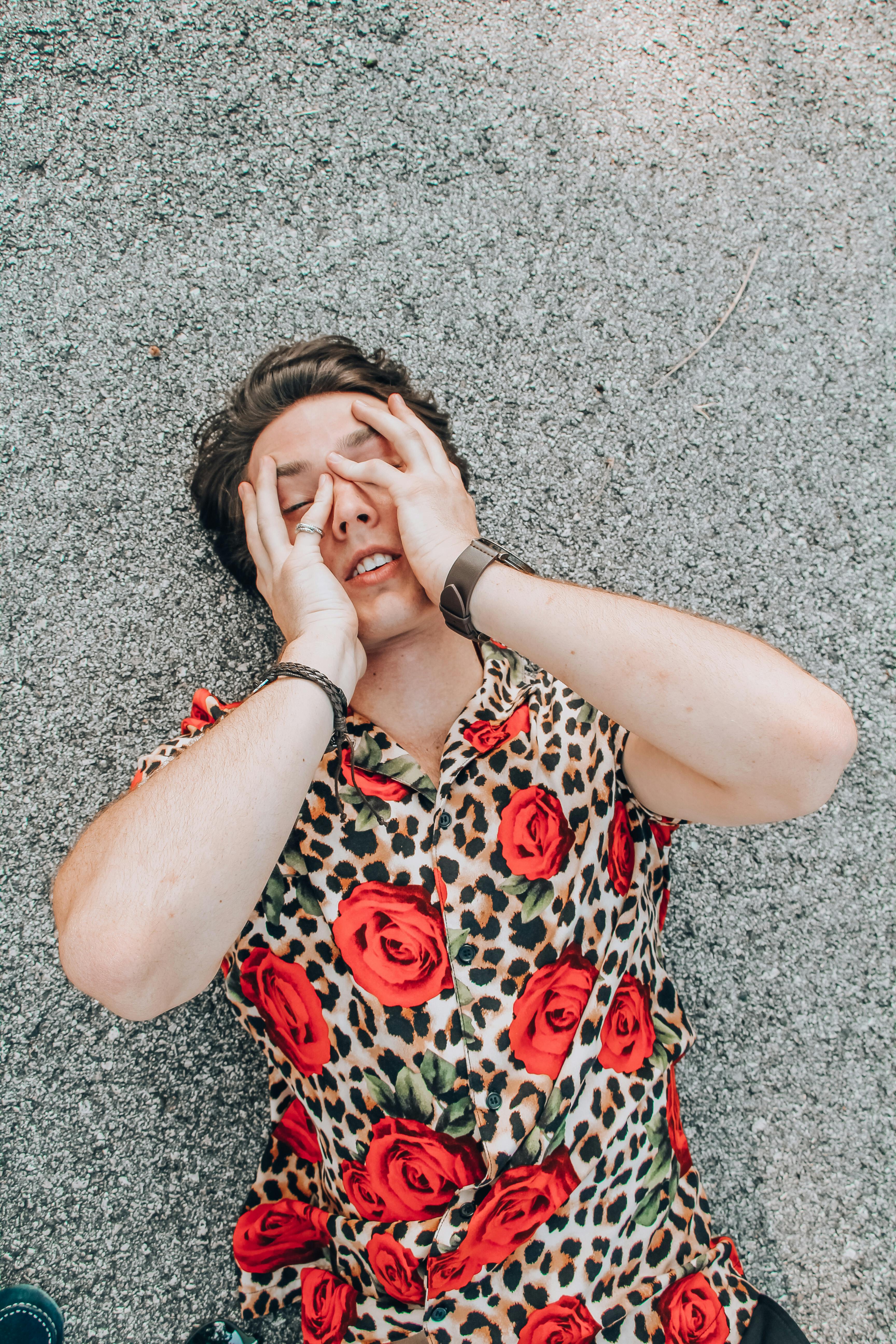 From above of young dreamy male in stylish apparel touching face with closed eyes while lying on roadway