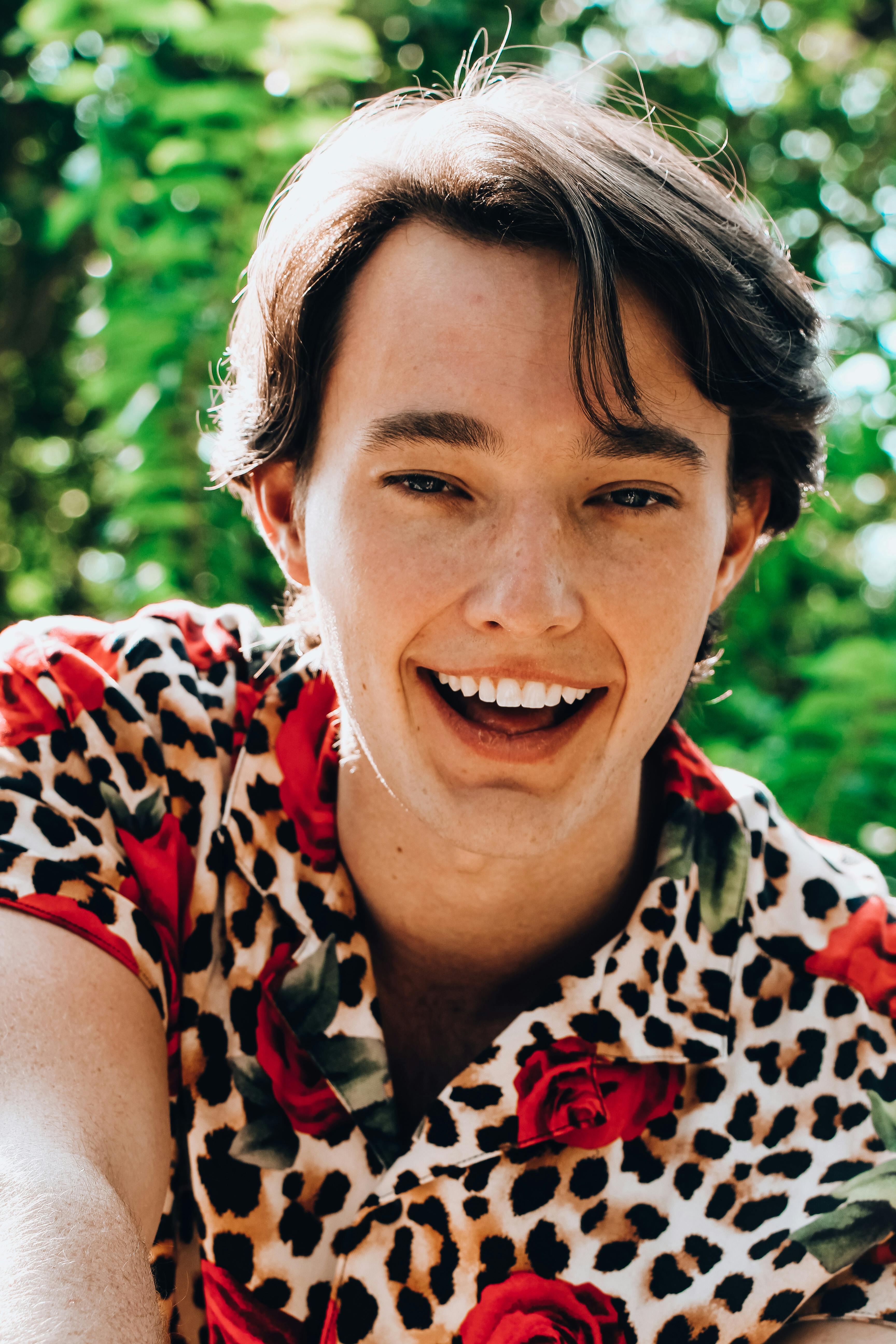 Excited young male in trendy shirt laughing and looking at camera contentedly while enjoying warm sunny day in nature