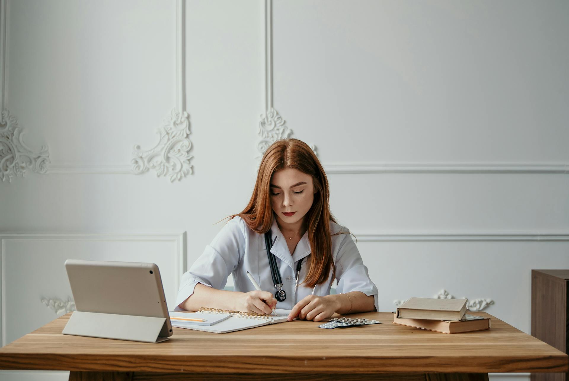 A woman doctor writing notes at a desk with a tablet and books, capturing a professional office setting.