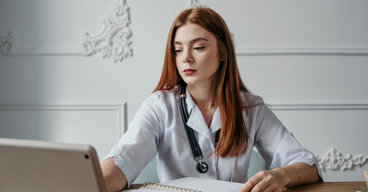 A professional female doctor wearing a stethoscope, working on a laptop at a desk with notebooks and medicines.