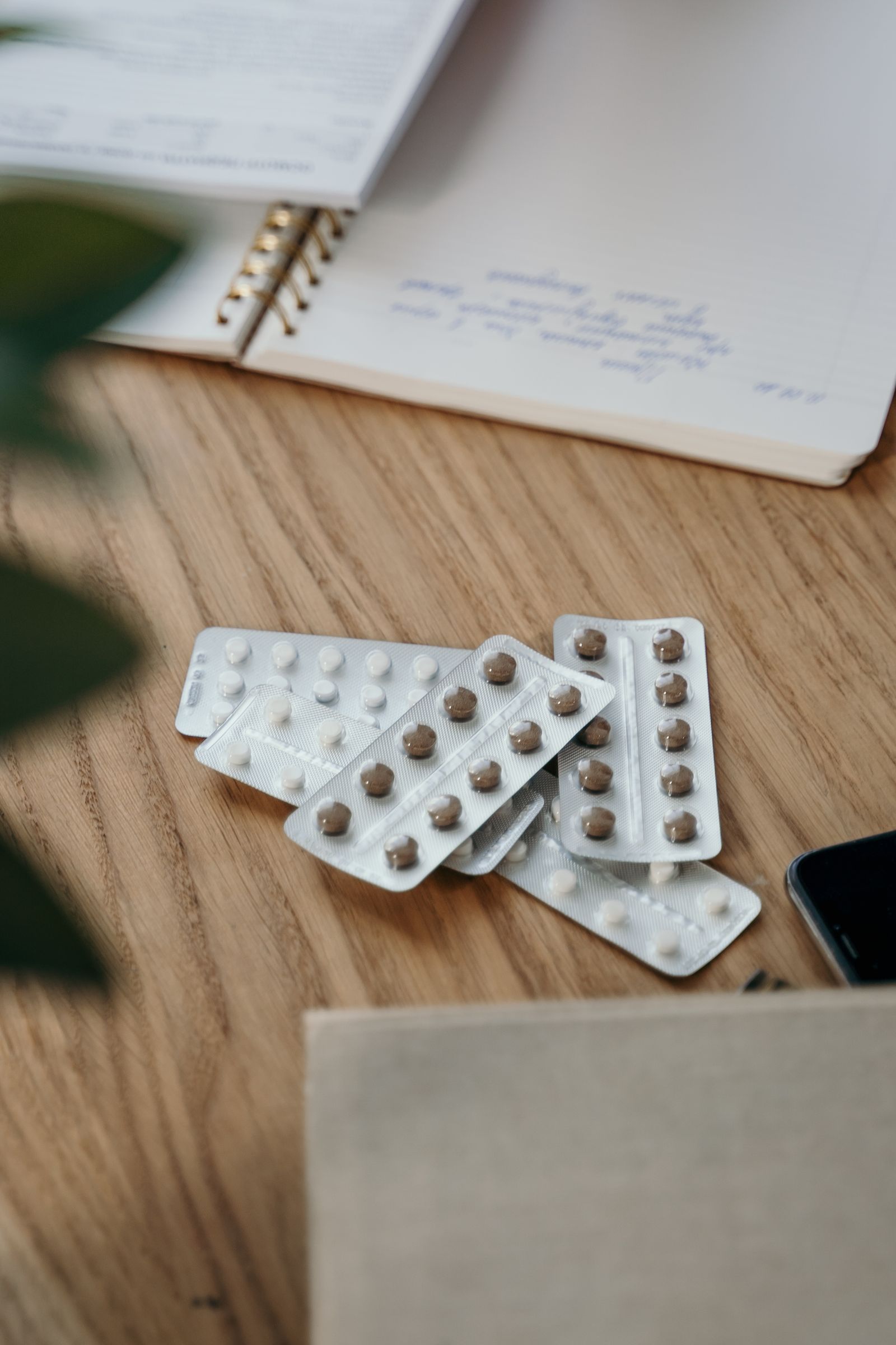 Blister packs of pills layered on a wooden tabletop beside an open spiral notebook with handwritten notes, soft natural light.