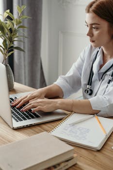 A professional female doctor typing on a laptop in her office with medical notes and stethoscope.