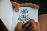 Crop anonymous talented person creating difficult mehendi sketch in album with white sheets