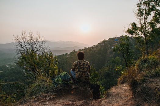 Back biew of anonymous male observing picturesque mountains and forest on cliff with trees in sunrise