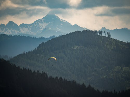 From above distant paraglider over wooded mountain slope under cloudy sky at daytime