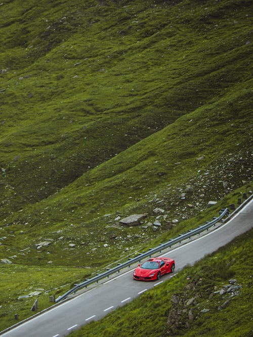 From above of red sports car driving along asphalt road in green grassy valley in daytime