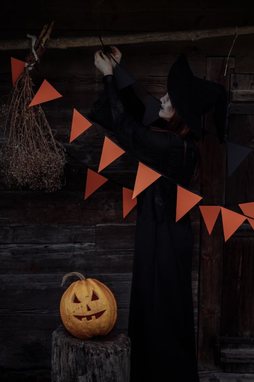 Free Witch in dark costume decorates woodshed with pumpkins and garlands for Halloween. Stock Photo