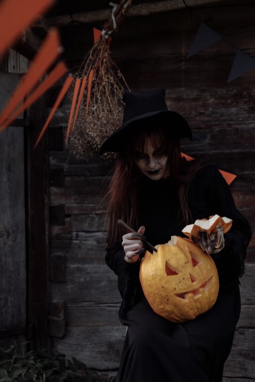 Free A witch in dark attire carves a jack-o'-lantern in a spooky, low-light setting. Stock Photo
