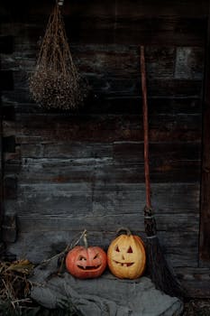 A creepy Halloween scene with carved pumpkins and a witch's broom against a wooden barn wall.