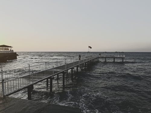 Black and white back view of unrecognizable travelers on fenced dock on foamy sea with horizon under sky