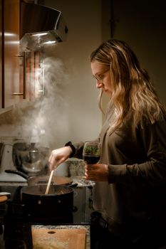 Adult woman cooking on a stove while holding a wine glass in a warm, cozy kitchen setting.