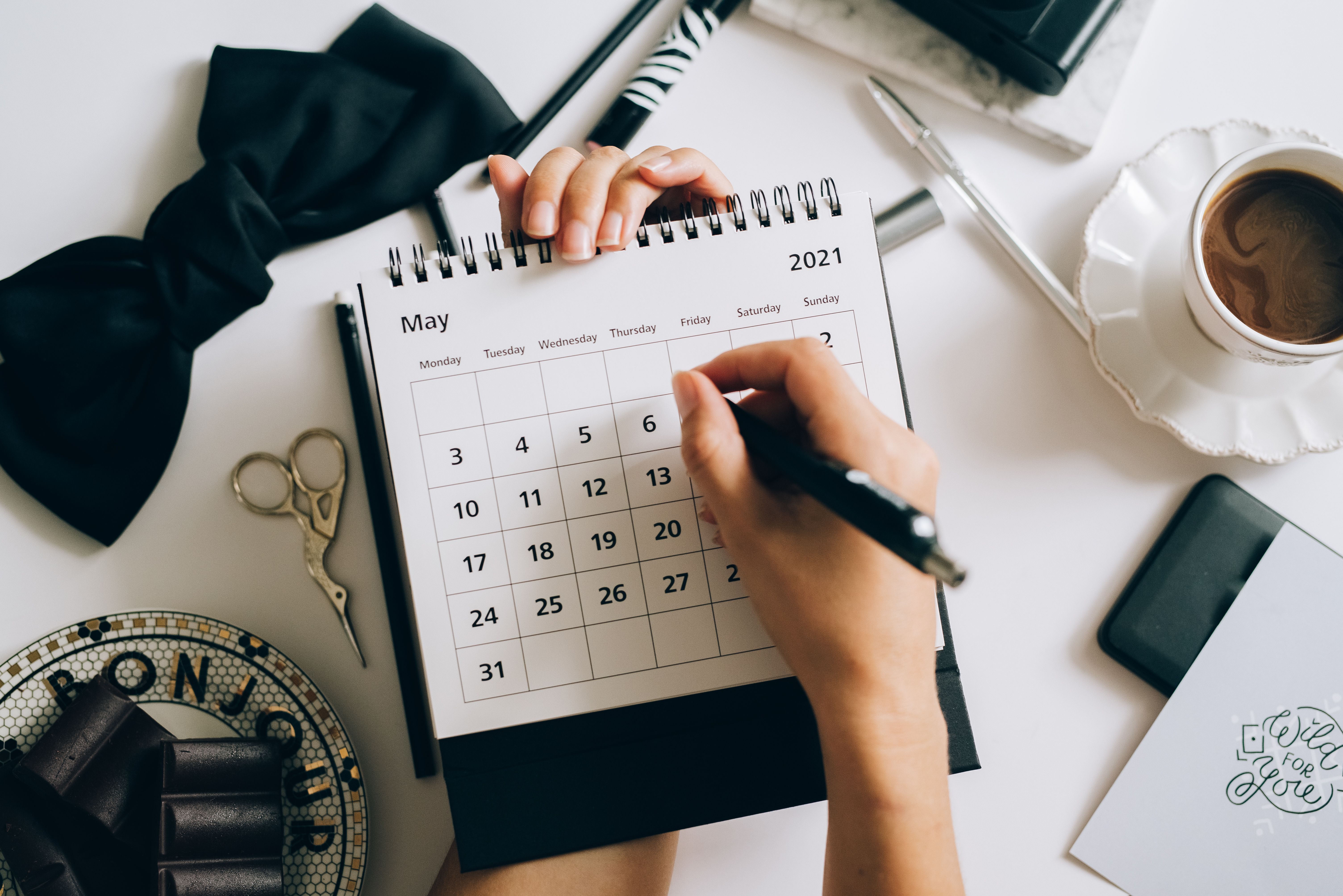 Person marking dates on a monthly calendar at a desk with stationery and a cup of coffee