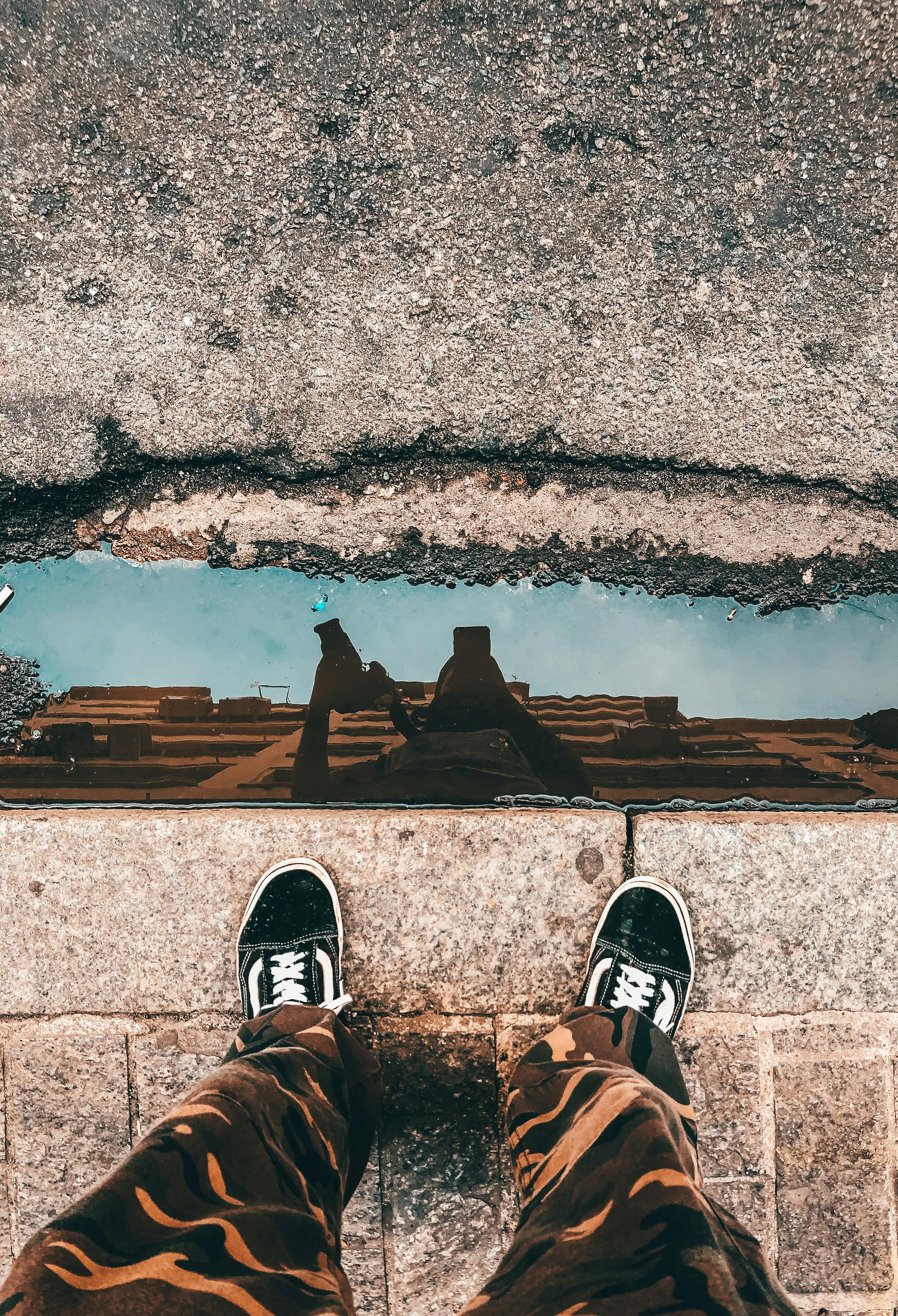 unrecognizable person standing on paved sidewalk near puddle reflecting building and blue sky