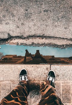 From above of crop anonymous person in trendy outfit taking selfie in puddle reflecting modern building and blue sky