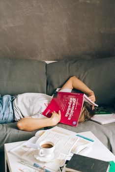 一名疲惫的学生躺在沙发上，脸上盖着一本书，周围散落着学习材料和咖啡