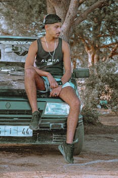 A man in casual sportswear sits on a parked car in a wooded area, enjoying leisure time.