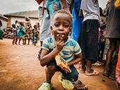 A young boy poses in an outdoor street setting surrounded by people, holding a snack, and showing a thoughtful expression.