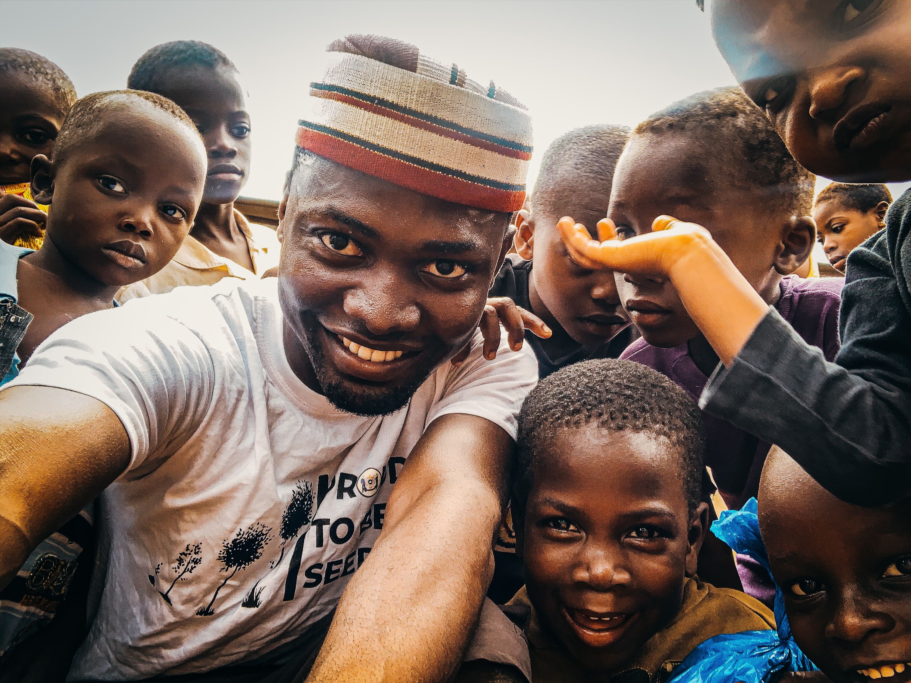 A cheerful group of African children and a man smiling while taking a selfie outdoors during the day.