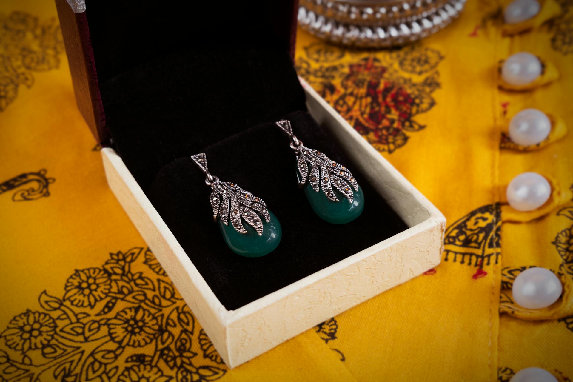 Close-up of silver and green gemstone earrings in a box on vibrant fabric.