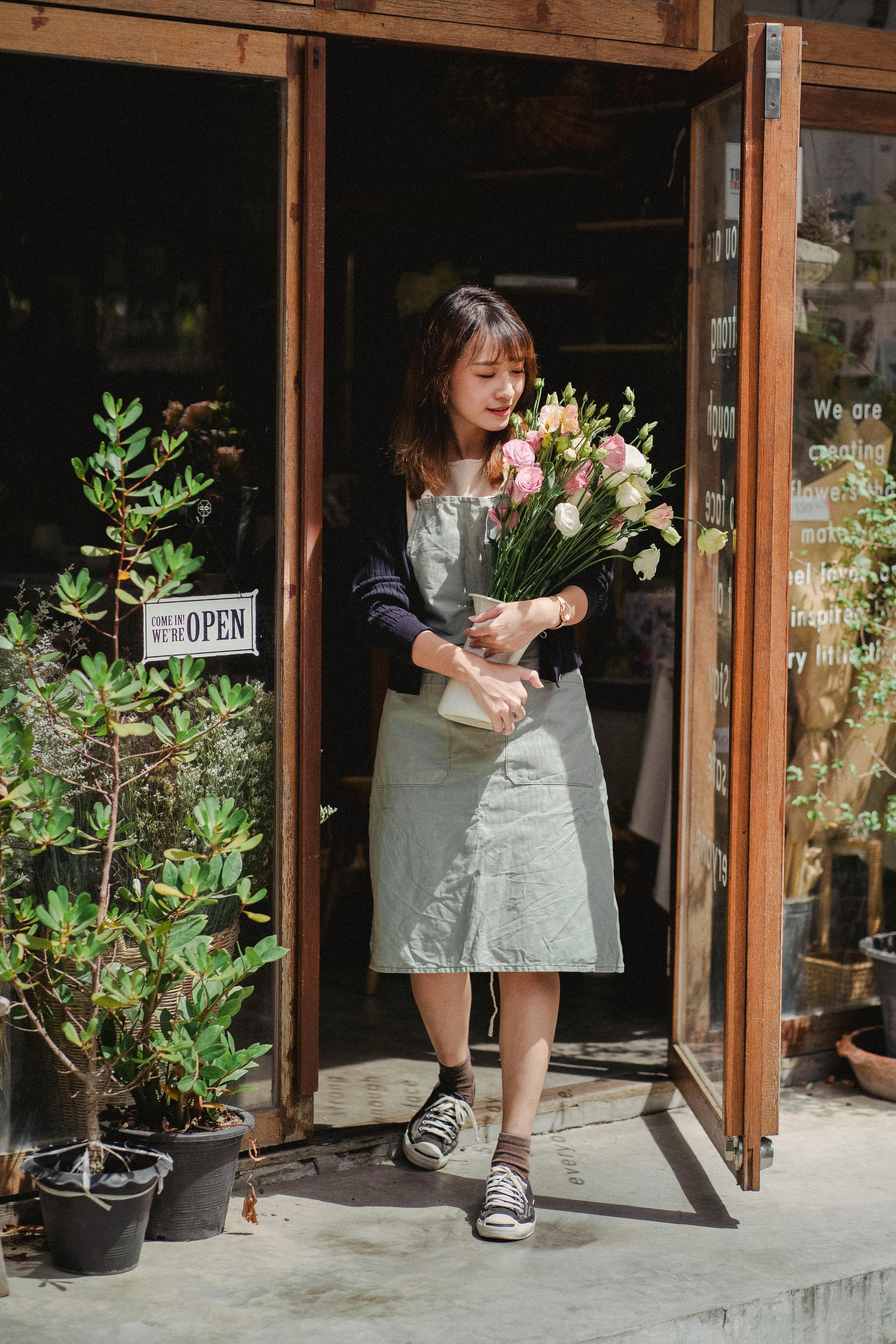 positive asian female florist standing with bouquet at shop doorway