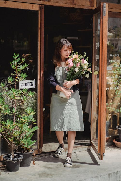 Free Positive Asian female florist standing with bouquet at shop doorway Stock Photo