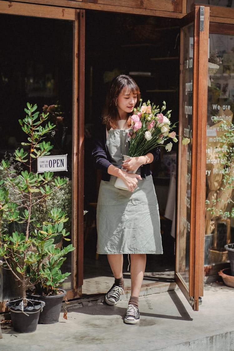 Positive Asian Female Florist Standing With Bouquet At Shop Doorway