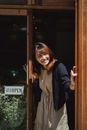 Positive smiling Asian female store worker opening store door and looking away contentedly