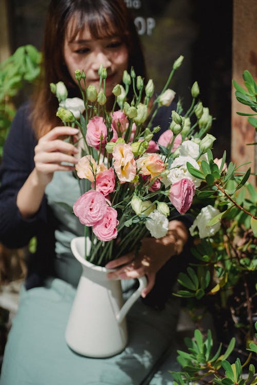 Crop positive Asian female florist in apron arranging blooming fragrant flowers in vase on sunny weather