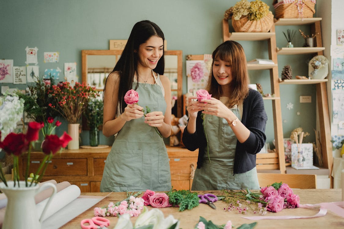 Two women in aprons smiling while arranging pink roses on a table in a flower shop.