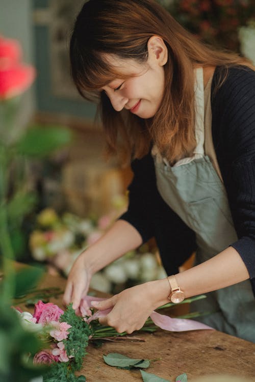 Side view of cheerful Asian female designer tying bouquet of fresh flowers in floral shop