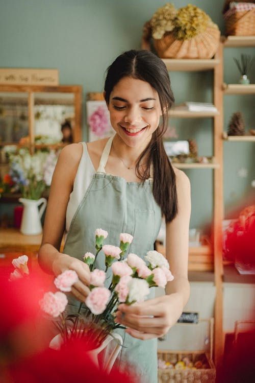 Smiling female florist in apron standing in flower shop and arranging flowers during work day