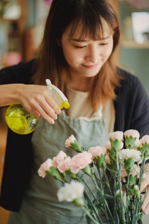 Young female florist in apron spraying fresh water on flowers during work in floral shop