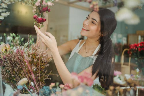 Young smiling dark haired female florist in apron arranging flowers while working in floral shop