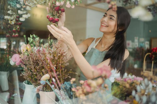 Smiling female florist in apron standing near table with flowers and making bouquets in floral shop