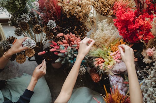 From above of crop faceless florists in aprons sitting near blooming flowers and arranging plants during work day