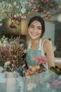 Through window of charming female worker in apron standing among flowers in store smiling at camera