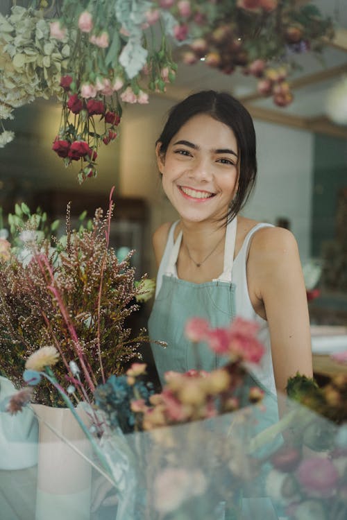 Free Through window of charming female worker in apron standing among flowers in store smiling at camera Stock Photo