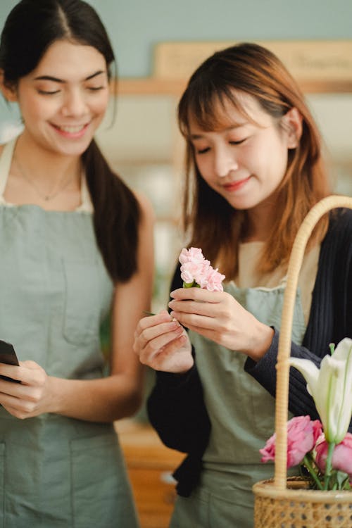 Happy multiethnic female florists wearing aprons and making composition of flowers while creating order during work in floral store with blurred background
