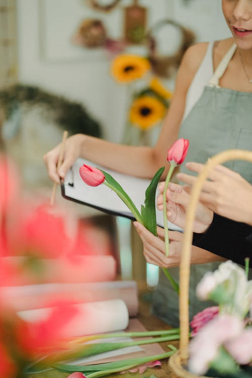 Unrecognizable female coworkers standing at table with tulips and writing notes on clipboard while working in florist atelier with blurred background