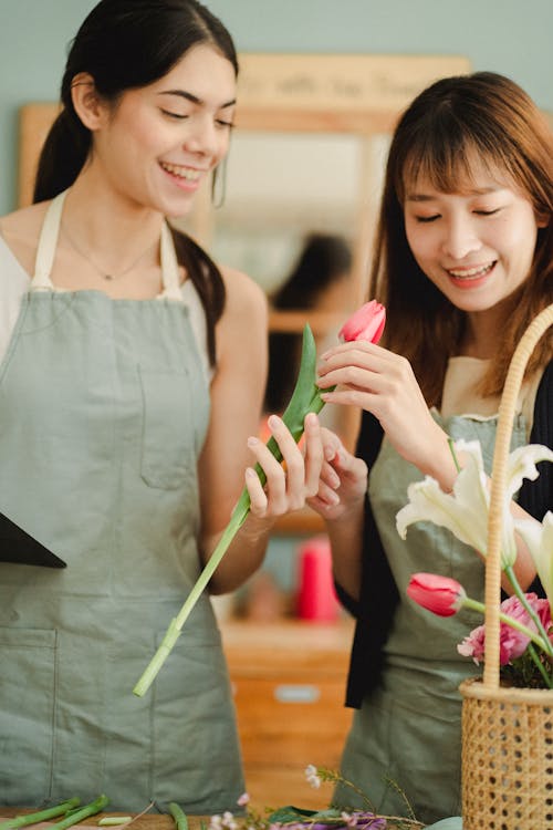 Positive multiethnic female coworkers wearing aprons while making composition of fresh tulips in wicker basket while working in florist shop