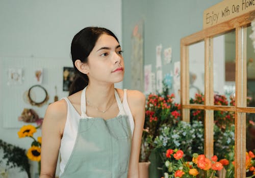 Free Wistful female florist with dark hair wearing apron standing near mirror with wooden frame and blooming flowers in floral store Stock Photo
